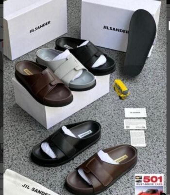 A display of Jil Sander two-strap leather slides in brown and black, accompanied by their signature boxes and packaging, embodying Modern Luxury and Urban Edge.