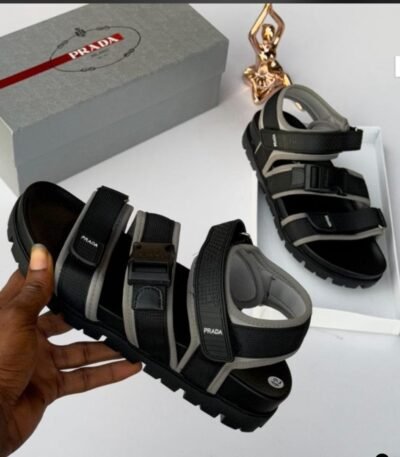 A pair of black and grey Prada sandals on a white background, with a hand holding one sandal and the other on a white box, next to a grey Prada box and a gold statue.