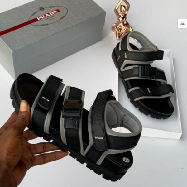 A pair of black and grey Prada sandals on a white background, with a hand holding one sandal and the other on a white box, next to a grey Prada box and a gold statue.