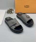 Grey UGG slide sandals with zigzag stitching and gold-tone buckle on a white surface, exuding Modern Luxury and Urban Edge.