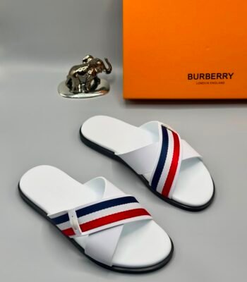 A pair of white Burberry slides with red and blue stripes, displayed with an orange Burberry box and a silver elephant figurine.