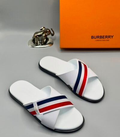 A pair of white Burberry slides with red and blue stripes, displayed with an orange Burberry box and a silver elephant figurine.