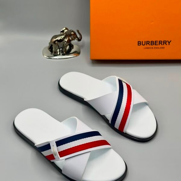 A pair of white Burberry slides with red and blue stripes, displayed with an orange Burberry box and a silver elephant figurine.