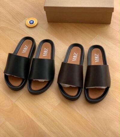 Two pairs of black leather slides with ZARA branding on the insoles, displayed on a wooden floor next to a brown shoe box and a small yellow container.