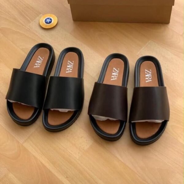 Two pairs of black leather slides with ZARA branding on the insoles, displayed on a wooden floor next to a brown shoe box and a small yellow container.