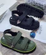 Luxury chunky sandals in black and green, featuring two wide straps with silver buckles, displayed on a wooden surface with Alexander McQueen packaging.