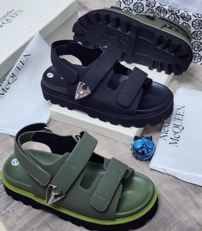 Luxury chunky sandals in black and green, featuring two wide straps with silver buckles, displayed on a wooden surface with Alexander McQueen packaging.