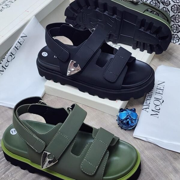 Luxury chunky sandals in black and green, featuring two wide straps with silver buckles, displayed on a wooden surface with Alexander McQueen packaging.