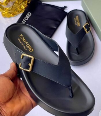 A pair of luxurious black slides with gold buckles, presented on a white surface with a black pouch and box in the background.