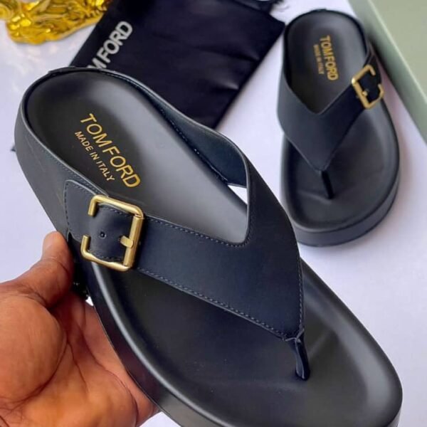 A pair of luxurious black slides with gold buckles, presented on a white surface with a black pouch and box in the background.
