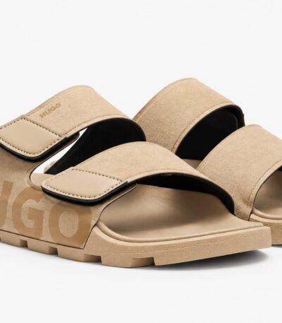 A pair of tan-colored HUGO slides with two wide straps and a chunky sole, set against a clean white background.