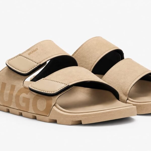 A pair of tan-colored HUGO slides with two wide straps and a chunky sole, set against a clean white background.