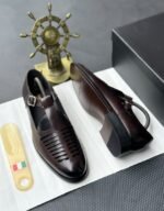 A pair of luxurious black leather monk strap shoes on a white cloth, accompanied by a shoe tree and a decorative ship's wheel, exuding modern luxury and urban edge.