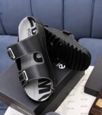 A pair of black leather slides with two adjustable straps and a thick, ridged sole, resting on a shoe box.