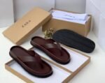 A pair of white leather slides with black outsole, presented in an open brown shoe box on a white surface.