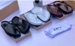 A pair of white leather slides with black outsole, presented in an open brown shoe box on a white surface.