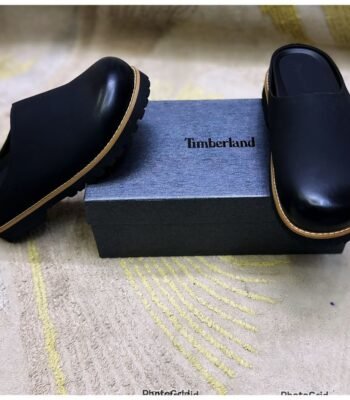 A pair of black leather mules with tan soles, positioned on either side of a black shoe box with the Timberland logo.