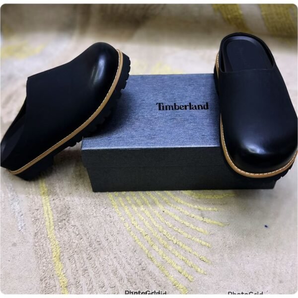 A pair of black leather mules with tan soles, positioned on either side of a black shoe box with the Timberland logo.