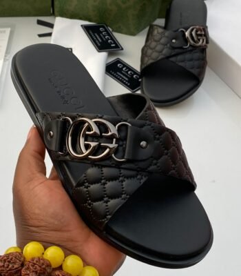 A pair of luxurious black Gucci slides with gold-tone buckle and quilted design, presented on a white surface with Gucci packaging in the background.