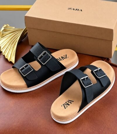A pair of black sandals with double silver buckles and tan insoles, placed on a brown leather surface next to a gold decorative item and a Zara shoe box.
