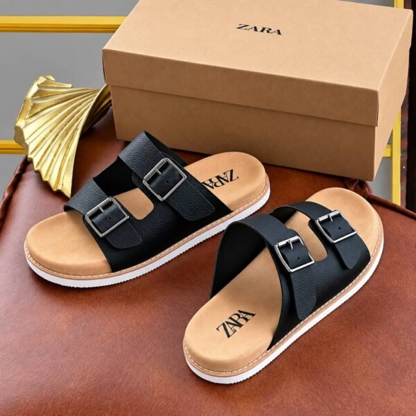A pair of black sandals with double silver buckles and tan insoles, placed on a brown leather surface next to a gold decorative item and a Zara shoe box.