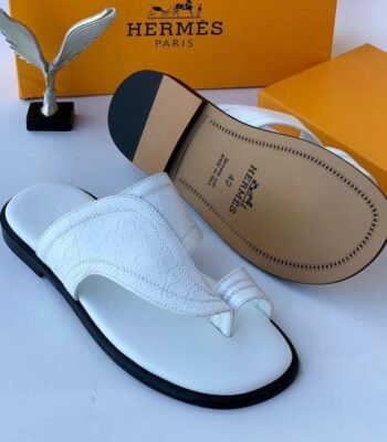 A pair of white leather slides with black soles, displayed with Hermes Paris branding and packaging, embodying Modern Luxury and Urban Edge.
