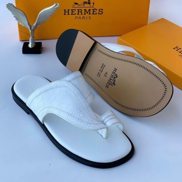 A pair of white leather slides with black soles, displayed with Hermes Paris branding and packaging, embodying Modern Luxury and Urban Edge.