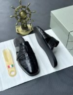A pair of black leather half shoes with crocodile-textured straps, displayed on a white surface with a gold Italian flag keychain and a decorative gold ship's wheel in the background.
