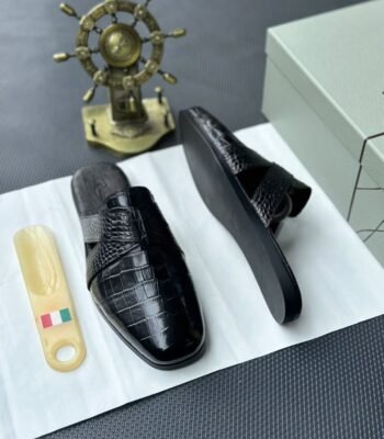 A pair of black leather half shoes with crocodile-textured straps, displayed on a white surface with a gold Italian flag keychain and a decorative gold ship's wheel in the background.