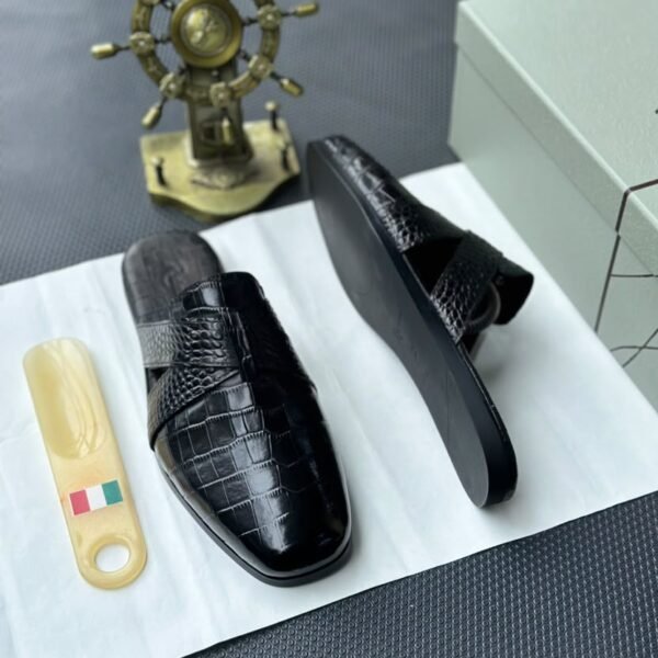 A pair of black leather half shoes with crocodile-textured straps, displayed on a white surface with a gold Italian flag keychain and a decorative gold ship's wheel in the background.