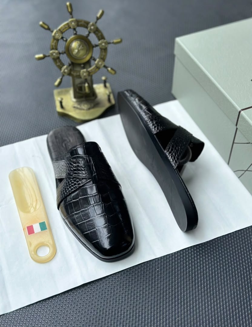 root A pair of black leather half shoes with crocodile-textured straps, displayed on a white surface with a gold Italian flag keychain and a decorative gold ship's wheel in the background.