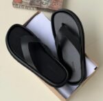 A pair of white leather slides with black outsole, presented in an open brown shoe box on a white surface.