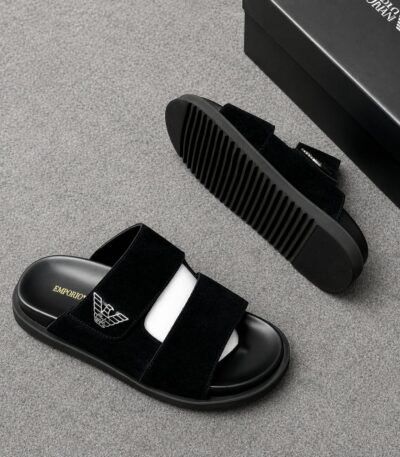 A pair of black Emporio Armani slides with a suede upper and ridged outsole, displayed on a grey surface with a black shoe box in the background.