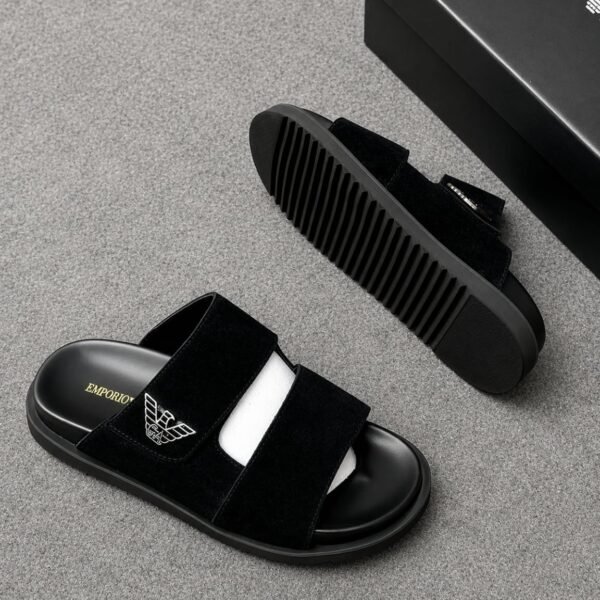 A pair of black Emporio Armani slides with a suede upper and ridged outsole, displayed on a grey surface with a black shoe box in the background.