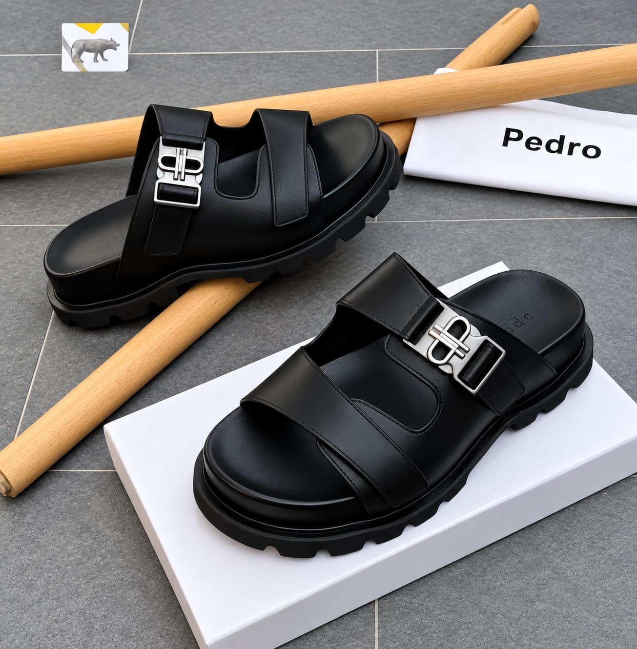 root A pair of black leather sandals with silver buckles, placed on a white shoe box and a grey tiled floor, exuding Modern Luxury and Urban Edge.