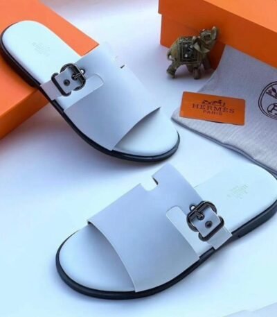 A pair of white slides with black buckles and soles, placed on a white surface with an orange box and Hermes Paris branding, embodying Modern Luxury and Urban Edge.