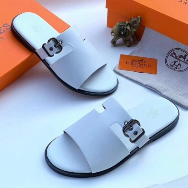 A pair of white slides with black buckles and soles, placed on a white surface with an orange box and Hermes Paris branding, embodying Modern Luxury and Urban Edge.