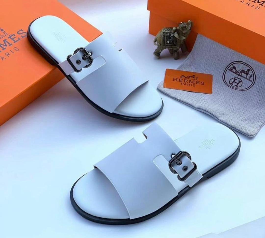root A pair of white slides with black buckles and soles, placed on a white surface with an orange box and Hermes Paris branding, embodying Modern Luxury and Urban Edge.