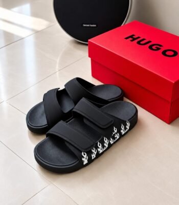 A pair of black slides with white flame details next to a red HUGO box and a black harman kardon speaker on a beige tiled floor.