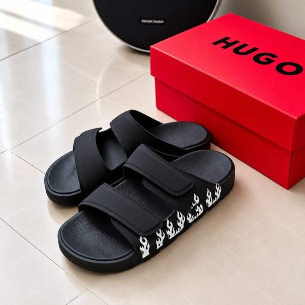 A pair of black slides with white flame details next to a red HUGO box and a black harman kardon speaker on a beige tiled floor.