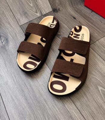 A pair of luxurious brown slides with bold lettering on the insoles, exuding Modern Luxury and Urban Edge on a sleek wooden floor.