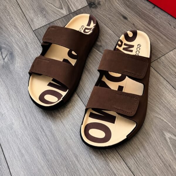 A pair of luxurious brown slides with bold lettering on the insoles, exuding Modern Luxury and Urban Edge on a sleek wooden floor.
