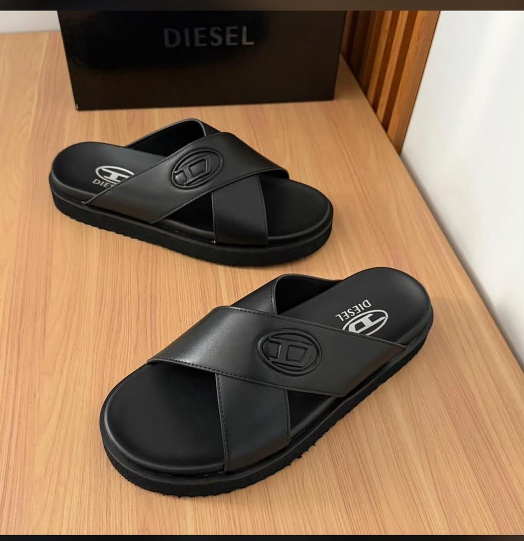 root A pair of black Diesel slides with white logo on a wooden table next to a black box.