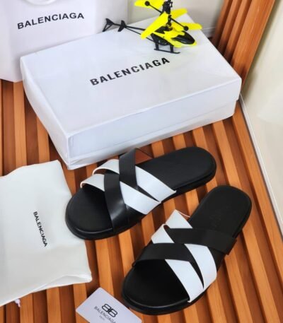 A pair of black and white Balenciaga slides on a wooden slatted surface, accompanied by a white shoe box, dust bag, and shopping bag, all bearing the Balenciaga logo.