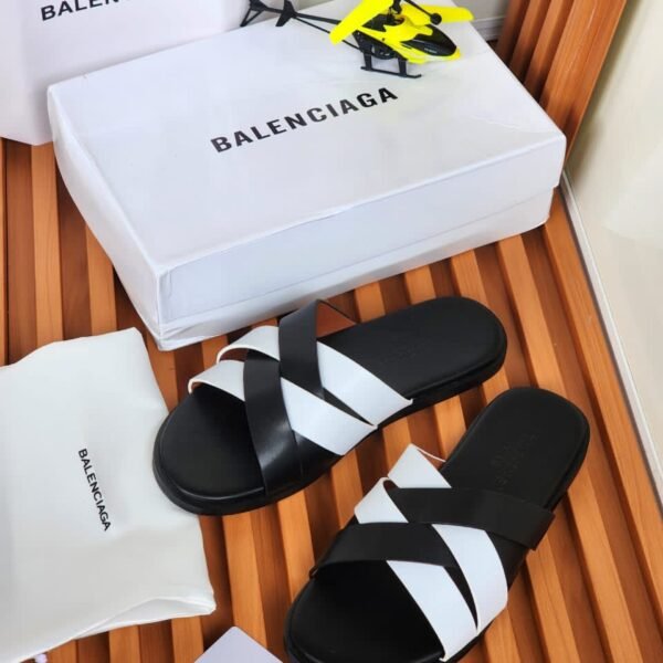 A pair of black and white Balenciaga slides on a wooden slatted surface, accompanied by a white shoe box, dust bag, and shopping bag, all bearing the Balenciaga logo.