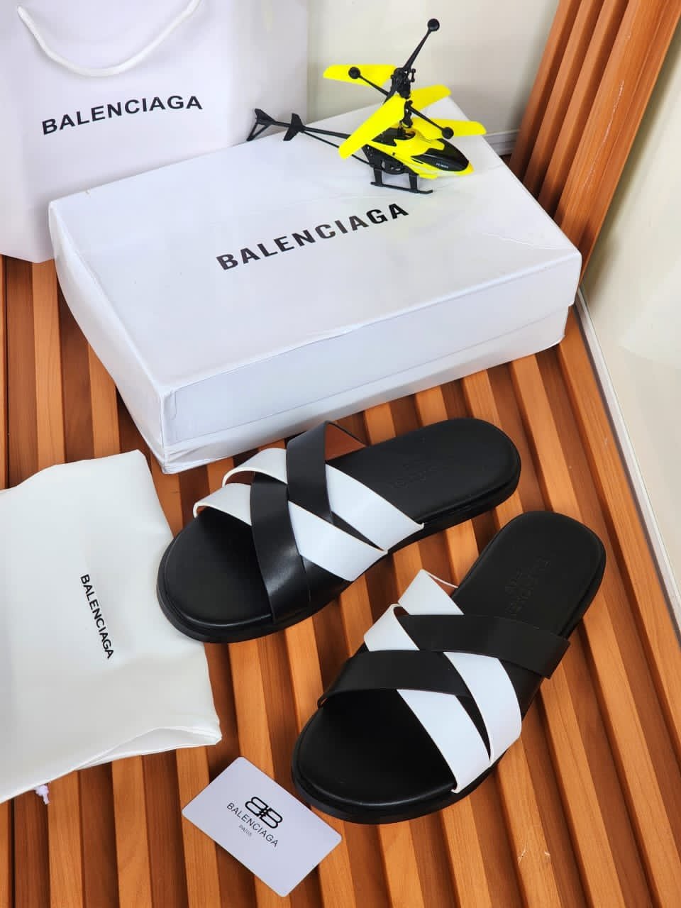 root A pair of black and white Balenciaga slides on a wooden slatted surface, accompanied by a white shoe box, dust bag, and shopping bag, all bearing the Balenciaga logo.
