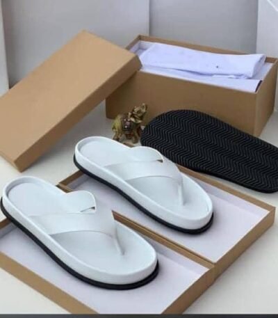 A pair of white leather slides with black outsole, presented in an open brown shoe box on a white surface.