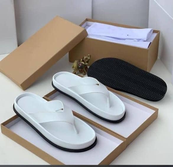 A pair of white leather slides with black outsole, presented in an open brown shoe box on a white surface.