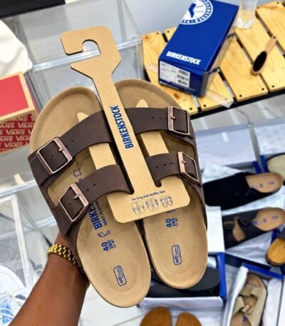 A pair of brown Birkenstock sandals with two adjustable buckles, presented on a cardboard holder, showcasing Modern Luxury and Urban Edge.