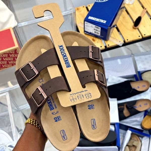 A pair of brown Birkenstock sandals with two adjustable buckles, presented on a cardboard holder, showcasing Modern Luxury and Urban Edge.